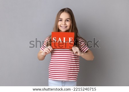 Portrait of little girl wearing striped T-shirt holding card with sale inscription, looking at camera with toothy smile. Indoor studio shot isolated on gray background.
