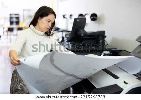 Woman using printer while working in print shop. Female printshop worker using printing device. Royalty-Free Stock Photo #2215268783