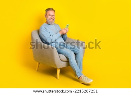 Full size photo of cheerful man sit chair hold use telephone chatting isolated on yellow color background