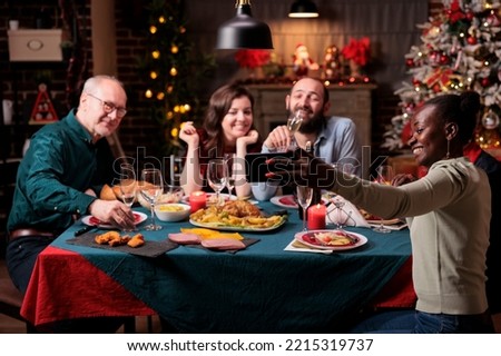 Woman taking family christmas celebration group photo on smartphone, friends portrait at festive dinner table. Diverse people gathering, celebrating xmas, eating traditional meal, drinking