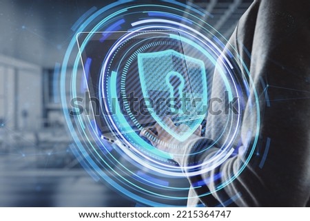 Close up of hacker hand using laptop with glowing shield hologram on blurry office interior background. Secure, data theft, malware, safety and web protection concept. Double exposure