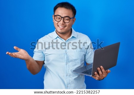 Chinese young man using computer laptop smiling cheerful presenting and pointing with palm of hand looking at the camera. 