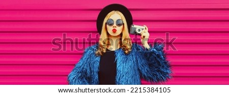 Portrait of stylish blonde young woman photographer with film camera taking picture and blowing her lips sends air kiss wearing blue fur coat, black round hat on pink background