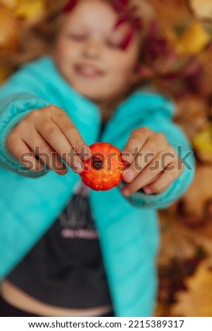Funny smiling girl lies on yellow leaves in the forest and holds small pumpkin in her hands. Pretty child enjoys the beautiful autumn nature.