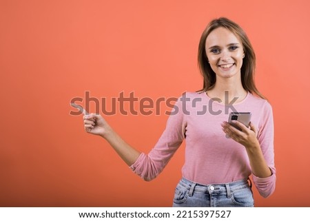 A young Caucasian girl in a pink jumper uses a mobile phone and points her finger at a copy place for advertising isolated on a red studio background.