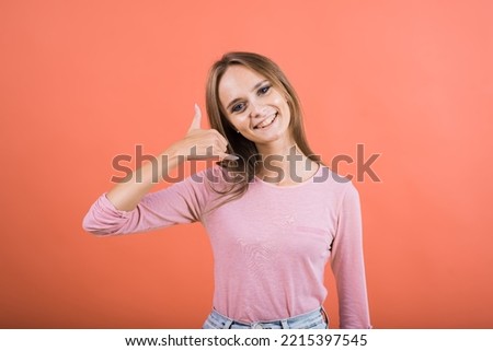 A young Caucasian girl in a pink jacket shows a hand and fingers gesture of a phone call isolated on a red studio background.