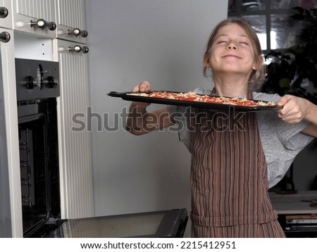 A teenager in an apron puts a baking sheet with pizza in the kitchen oven for cooking.Healthy home cooking.Leisure and hobby of schoolchildren.