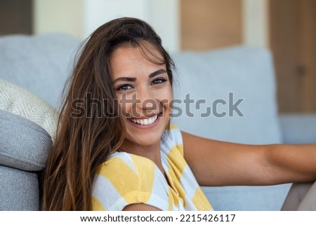 Happy young woman sitting on sofa at home and looking at camera. Portrait of beautiful woman smiling and relaxing during summer.