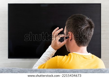 a man sticks headphones in his ear against the background of a TV screen.. watching TV with headphones at night.