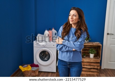 Young hispanic woman smiling confident standing with arms crossed gesture at laundry room