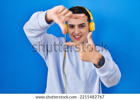 Non binary person listening to music using headphones smiling making frame with hands and fingers with happy face. creativity and photography concept. 