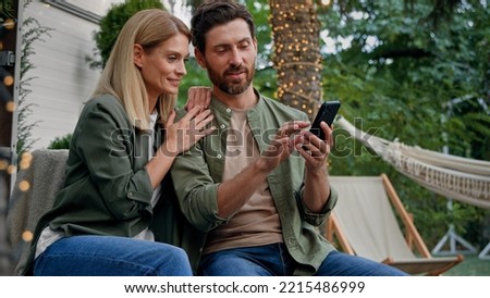 Married couple husband and wife Caucasian bearded man and blond woman 40s spouses outdoor in backyard at campsite looking at phone choose buy online booking with smartphone using mobile e-shopping app