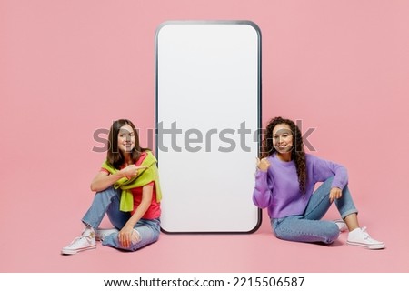 Full body young two friends women wears green purple shirts point finger on big huge blank screen mobile cell phone smartphone with workspace area isolated on pastel plain light pink color background