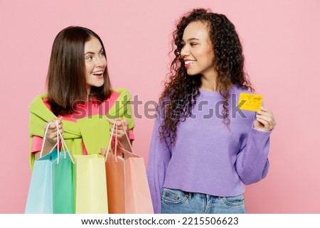 Young two friends women wears shirts hat hold in hand paper package bags after shopping together use credit bank card isolated on plain light pink color background. Black Friday sale buy day concept