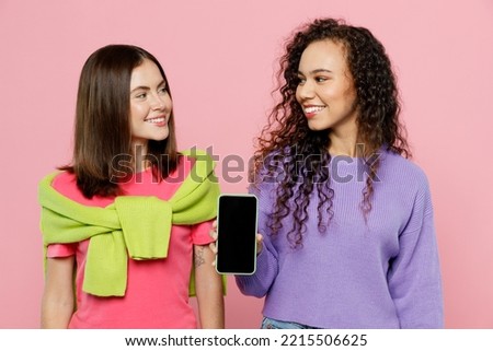 Young two friends women wears green purple shirts together hold in hand use mobile cell phone with blank screen workspace area look to each other isolated on pastel plain light pink color background