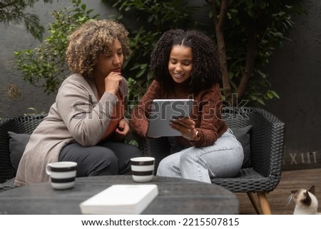 An African-American woman and her daughter use a digital tablet for online shopping. They sit on the patio and drink a coffee. The daughter is petting her siamese cat.