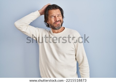 Handsome middle age man wearing casual sweater over blue background confuse and wonder about question. uncertain with doubt, thinking with hand on head. pensive concept. 
