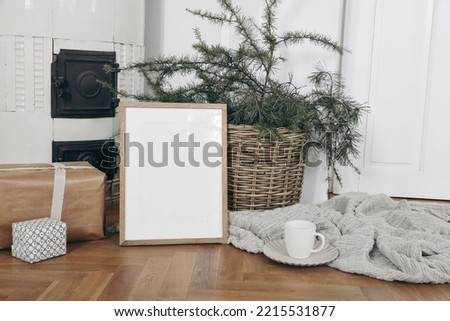 Festive Christmas indoor decor. Vertical wooden picture frame mockup on parquette floor. Pine, larch tree branches in basket, gift boxes. Cup of coffee. Old white tiled stove background.Empty template