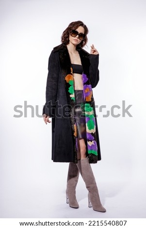 High fashion photo of a beautiful elegant young woman in a pretty black fur coat, suede boots, stylish sunglasses posing over white background. Make up, hairstyle. Slim figure.