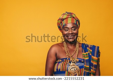 Smiling Ghanaian woman wearing a traditional attire against a yellow background. Happy black woman dressed in colourful Kente cloth and golden jewellery. Mature elegant woman embracing her rich cultur Royalty-Free Stock Photo #2215552231