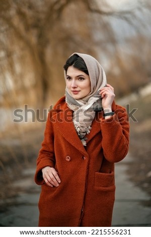 Young brunette girl in orange coat and woolen scarf straightens her headscarf with her hand, looking away, standing against backdrop of autumn park in blur.