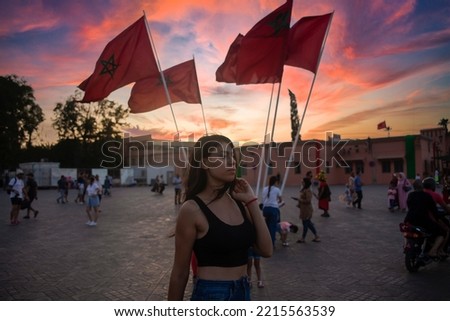 Beautiful Young tourist in Jemaa el Fna square in Marrakech next to the Moroccan flag at sunset, this city is very popular in Morocco and is very beautiful at sunset under an orange sky.
