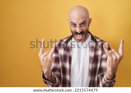 Hispanic man with beard standing over yellow background shouting with crazy expression doing rock symbol with hands up. music star. heavy concept. 