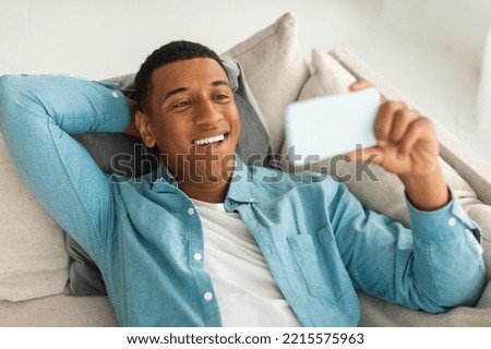 Happy millennial african american guy have video call, watching funny video on smartphone in free time in living room interior. Rest and relax at home with device, new app during covid-19 quarantine
