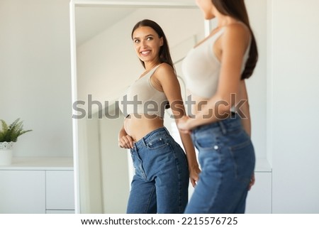Cheerful Lady Posing Wearing Skinny Jeans Smiling To Her Reflection In Mirror Standing After Successful Weight Loss At Home. Female Beauty And Style, Self Confidence Concept. Selective Focus Royalty-Free Stock Photo #2215576725