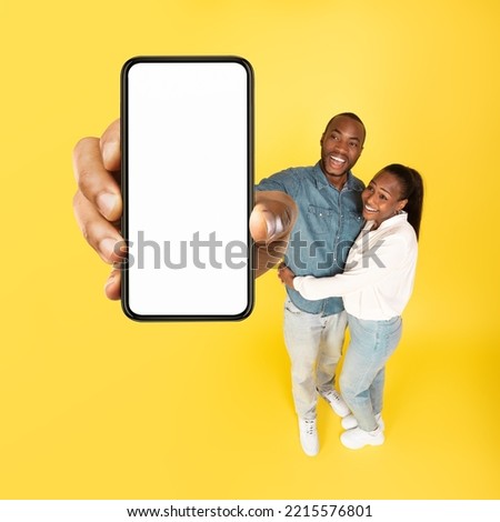 Cheerful Couple Showing Large Smartphone With Empty Screen Advertising Mobile Offer Smiling To Camera Posing Standing On Yellow Studio Background, High Angle Shot. Great Application. Square
