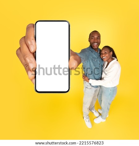 Cheerful Black Spouses Showing Huge Smartphone With Empty Screen To Camera Advertising Mobile Offer Posing Over Yellow Studio Background. Wow Application. Mockup, Square Shot