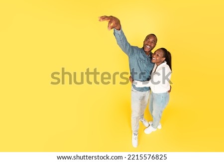 Look At This. Happy African American Spouses Advertising Something Holding Invisible Object In Fingers Posing Standing Near Copy Space Over Yellow Background. Studio Shot