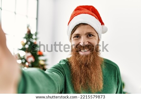 Young redhead man make selfie by the camera standing by christmas tree at home