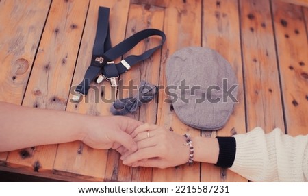 Baby boy is coming: a couple is holding hands, there are little boy's gentleman's clothes next to them on a wooden bench, a cap, a bow tie and a bridle
