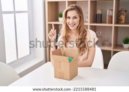 Beautiful blonde woman with cardboard box smiling with an idea or question pointing finger up with happy face, number one 