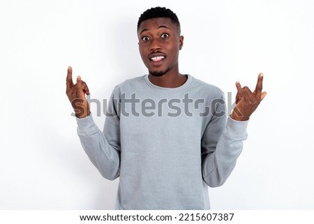 young handsome man wearing grey sweater over white background  makes rock n roll sign looks self confident and cheerful enjoys cool music at party. Body language concept.