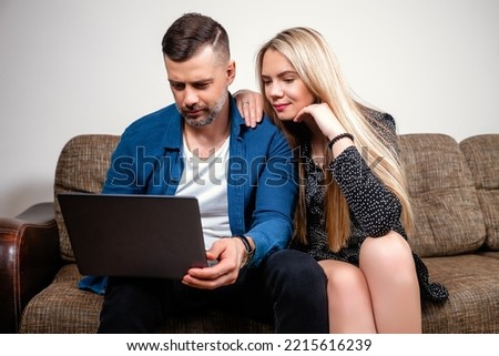 Pleasant married couple is sitting on the couch in the modern room and looking at the laptop screen. Happy young mixed race married spouse while browsing the web, shopping online or booking airline Royalty-Free Stock Photo #2215616239