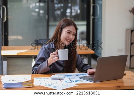 businesswoman holds a coffee cup before analyzing company sales using her laptop computer and documents on her desk.