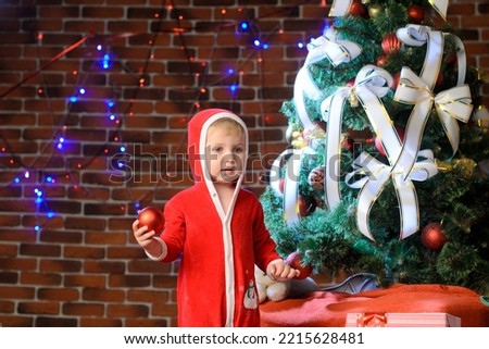 A little boy dressed as Santa Claus stands near a Christmas tree against a brick wall with bokeh