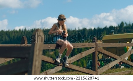 Young photographer wiping digital camera lens by t-shirt, sitting on wooden fence. Girl shooting beautiful nature landscape, concentrating on process, spending leisure time outdoor. Slow motion.