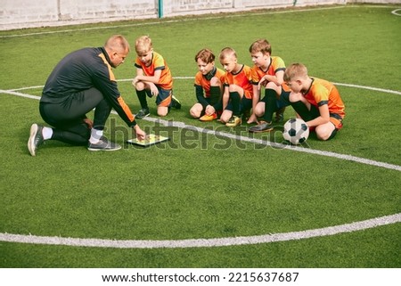 Football training. Soccer coach explaining game rules and strategy using tablet, map. Sports junior team sitting on grass pitch with trainer. Concept of sport, achievements, studying, goals, skills Royalty-Free Stock Photo #2215637687