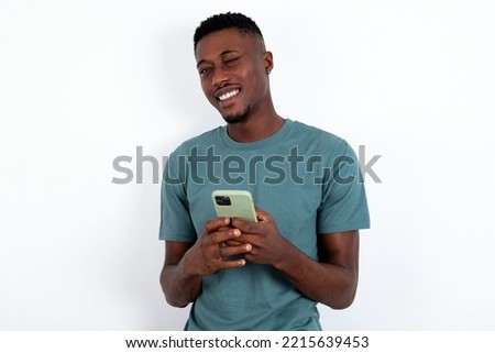 Pleased young handsome man wearing green T-shirt over white background using self phone and looking and winking at the camera. Flirt and coquettish concept.