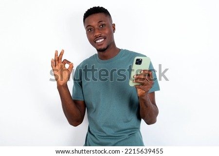 Happy young handsome man wearing green T-shirt over white background sending a message on his smartphone or taking a selfie  and making ok sign with his hand.