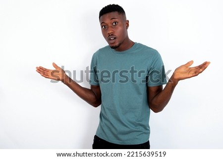 So what? Portrait of arrogant young handsome man wearing green T-shirt over white background shrugging hands sideways smiling gasping indifferent, telling something obvious. Royalty-Free Stock Photo #2215639519
