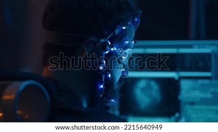 Back view of a brunette guy wearing futuristic headset with LED lights attached programs and develops code using multiple computer screen. Cyperpunk style. Sci-fi background. Neon lights.