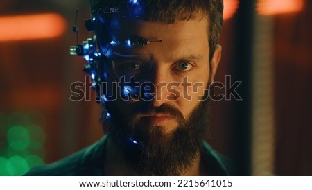 Brunette guy with dark beard wearing futuristic headset with one-eyed glasses, mic and ear piece looks at the camera with intense facial expressions. Neon lights in the background. Cyberpunk.