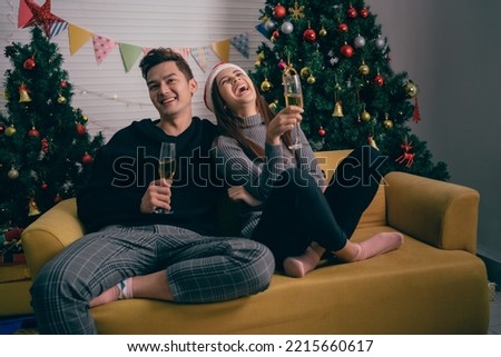 Happy Asian couple toasting champagne together, sitting on the sofa in the evening with a Christmas tree and lights in the background. Boyfriend and girlfriend at Christmas eve night