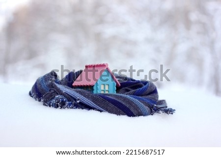 A model of a rustic house wrapped in a warm scarf on a blurry winter background. A toy cottage in the snow.