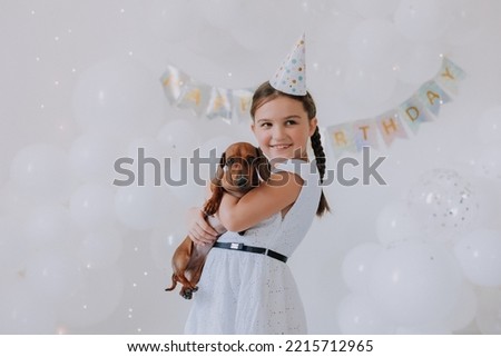 little girl with two pigtails in a white dress with her beloved dog dachshund in her arms celebrates her birthday. party with white balloons. High quality photo