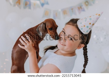 little girl with two pigtails in a white dress with her beloved dog dachshund in her arms celebrates her birthday. party with white balloons. High quality photo
